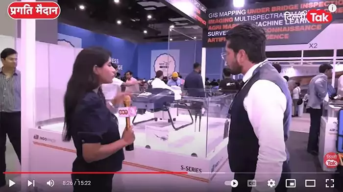 scene of exhibition where a man in black suit give an interview to a girl in black
                         dress while a few people are milling around them